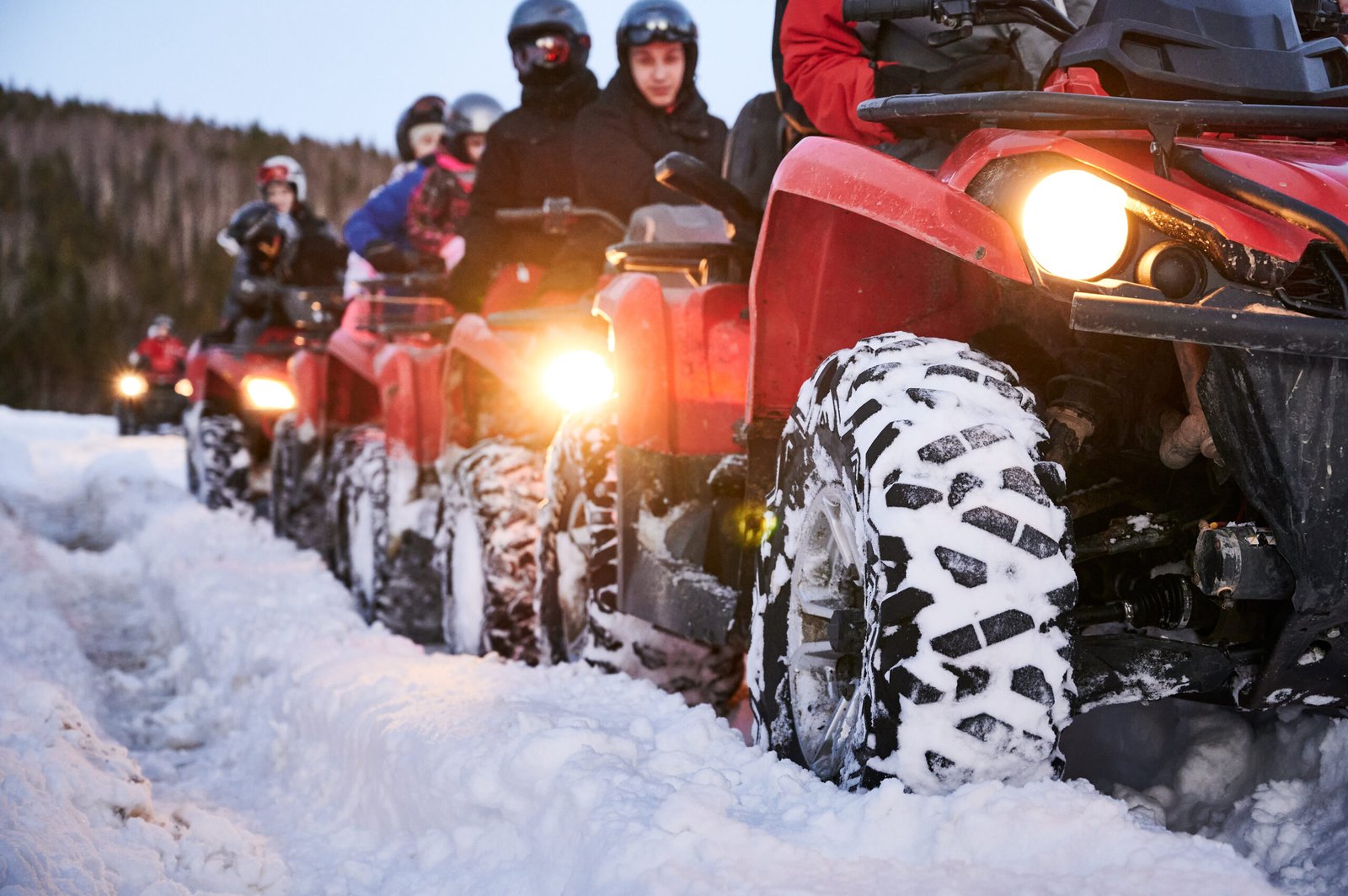 Focus on the wheel. People riding red all-terrain vehicles down snowy hill. Quad riders driving quad bikes with black snowy wheels on snow-covered trail. Concept of winter activities and quad biking.